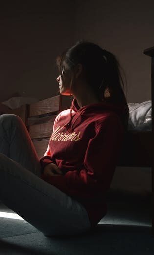 pexels-photo-3007355-3007355 Contemplative young woman in hoodie sitting on the floor in dimly lit room, symbolizing solitude and introspection.