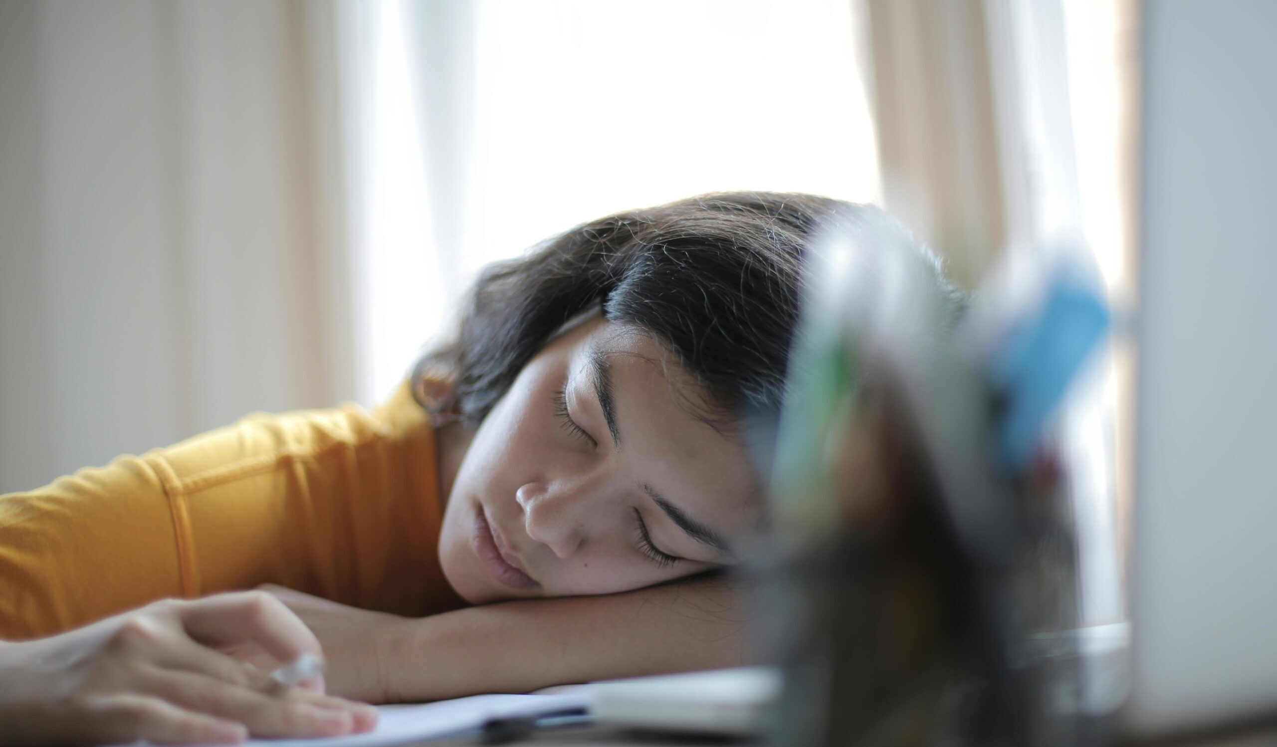 Young woman in yellow shirt resting her head on a desk, showing signs of fatigue and relaxation indoors.